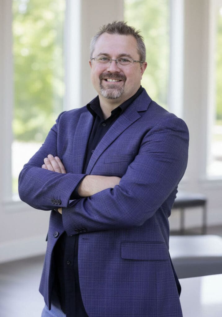 Professional portrait of Don Enos, Compass Real Estate agent in Bellingham, standing with arms crossed and smiling in a bright office setting.